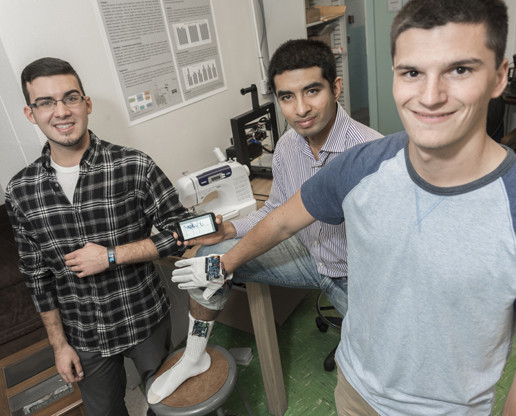 URI students join Assistant Professor of Biomedical Engineering Kunal Mankodiya, while displaying smart textiles, wearable items embedded with sensors, electronics and software that can collect data from patients, even though they are at home, and deliver it to doctors. From left: Nicholas Peltier, senior computer engineering major of Coventry, Professor Mankodiya, and Matt Constant, junior computer engineering major of West Warwick. URI Photo by Michael Salerno Photography.